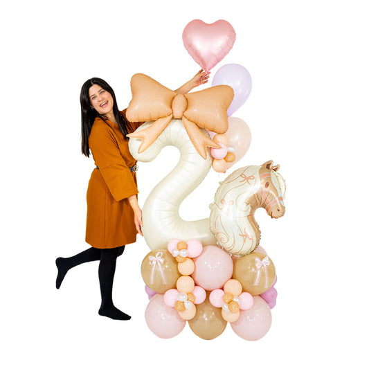 Woman standing beside a coquette horse birthday balloon arrangement with a cream number 2, pony foil, oversized peach bow and pastel balloon base.