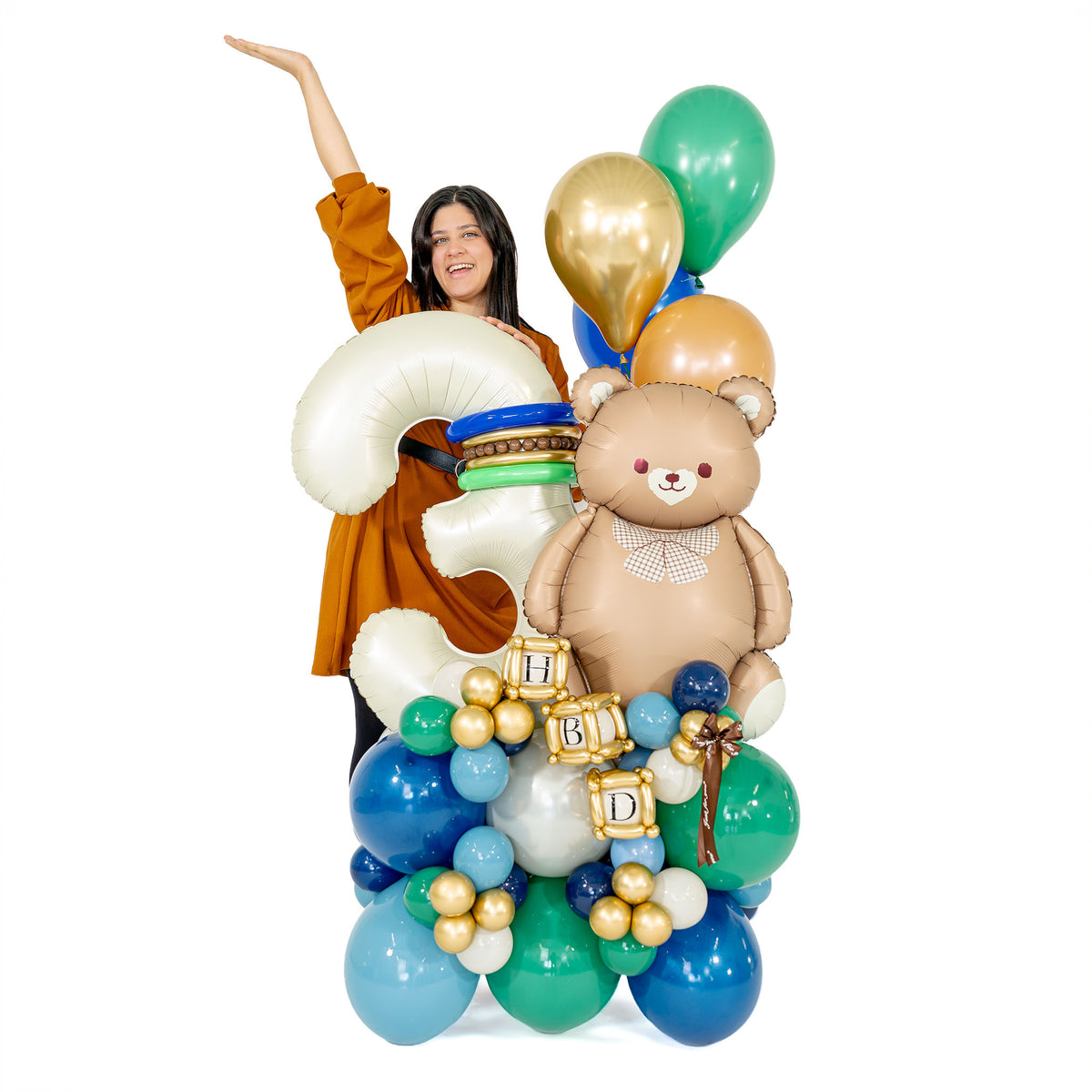 Woman standing behind a large teddy bear birthday balloon arrangement with a cream number 3 and blue, green and gold balloons.