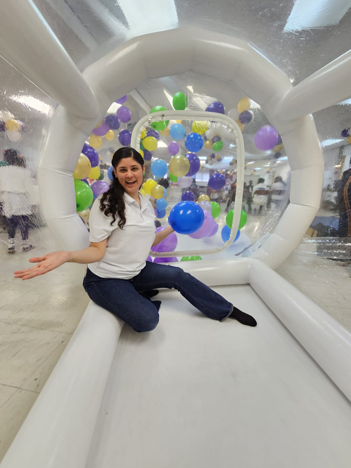 Woman sitting inside a transparent inflatable bubble with colorful balloons in an indoor setting.