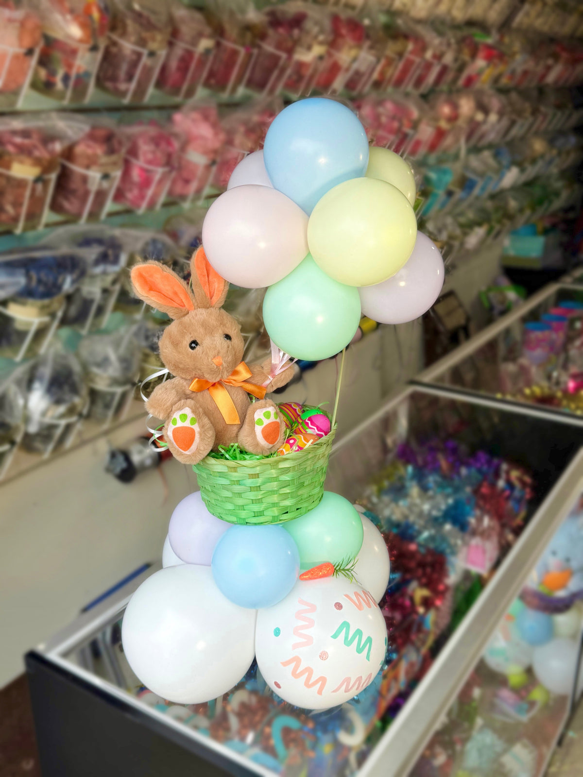Decorative arrangement of pastel balloons with a teddy bear and basket in a store setting.