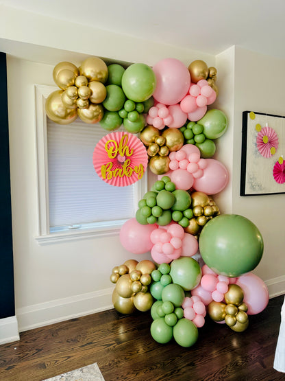 Balloon arch with gold, pink, and green balloons in a room setting.