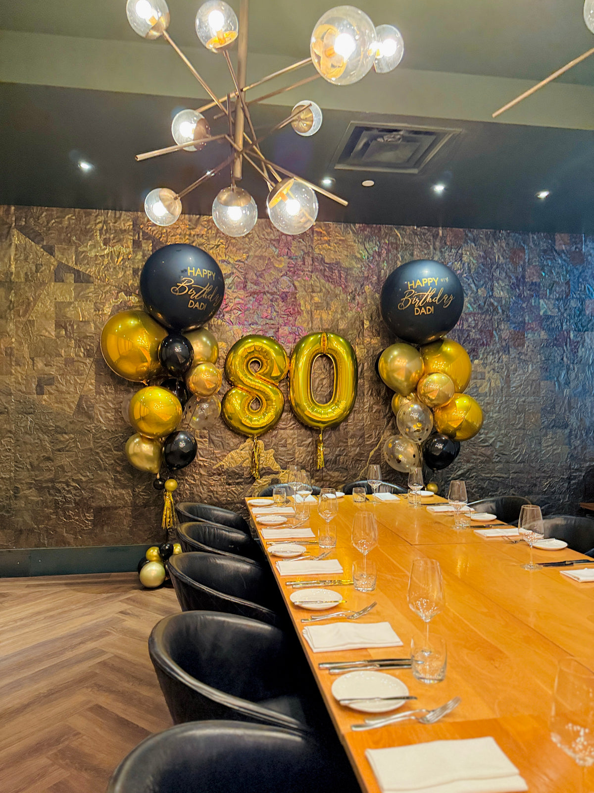 Dining room with a table set for a celebration, featuring gold and black balloons with 'Happy Birthday' and number '80'.