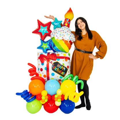 Woman beside a colourful cupcake birthday balloon arrangement with foil stars, a gift-box balloon and a rainbow base, showing its size